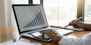 A person calculating rental property ROI with a calculator, laptop, and a rising bar graph.